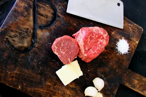 Picture of beef surrounded by herbs on a cutting board and a meat cleaver