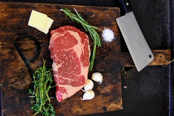 Picture of beef surrounded by herbs, and cheese on a cutting board, with a meat cleaver